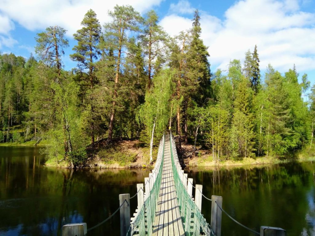 Aurinkoisessa säässä kylpevän, tyynen Harrisuvannon ylitse kulkee riippusilta kohden vastapäistä puiden peittämää saareketta. 