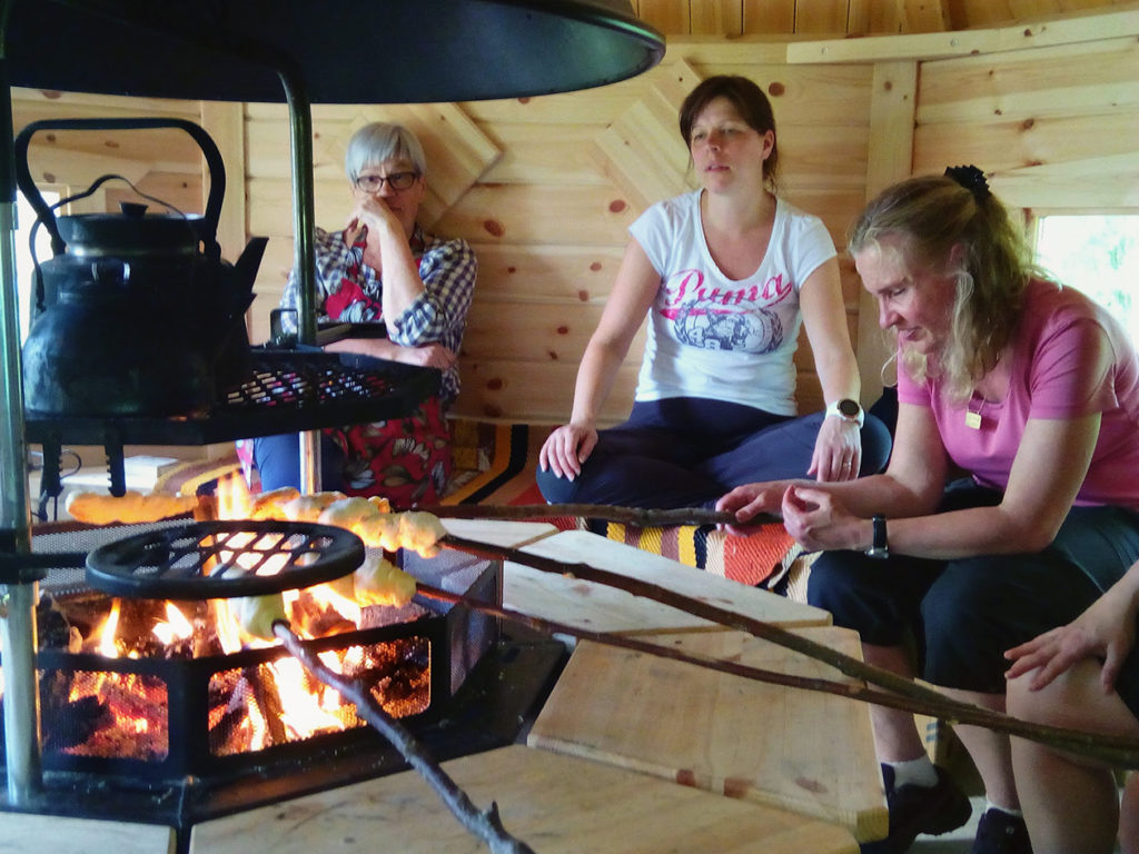 Retkeilijät rentoutumassa kodassa tulen lämmössä. Tikkupullat paistuvat ja vesi lämpenee nokipannussa tulen yllä. 