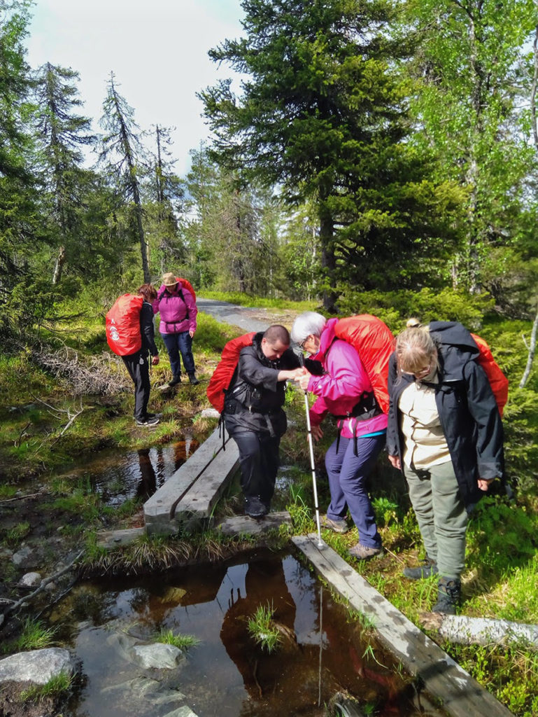 Lyhyt mutta vaativa kosteikon ylitys. Alkumatkasta kaksi puuta vieretysten, sitten tasapainottelua pyöreän tukin päällä ja askellus sivuun yhden laudan levyiselle pitkokselle. Anneli auttaa Outia pyöreän tukin yli.  Taija testaa maaston märkyyttä etualalla. Sirpa opastaa Heidiä ylityksen alkupäässä pienen kostean rinteen alastulossa. Vesi ei ole syvää mutta takuuvarmasti kylmää eikä syvyyttä tee mieli testata. Vedestä pilkistää lisäksi petollisia kiviä. 