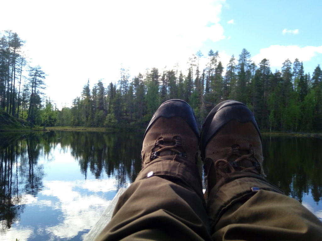 Vaelluskenkien peittämät jalat laiturilla lammen yllä. Valkoiset pilvet ja sininen taivas heijastuvat lammen pintaan. 