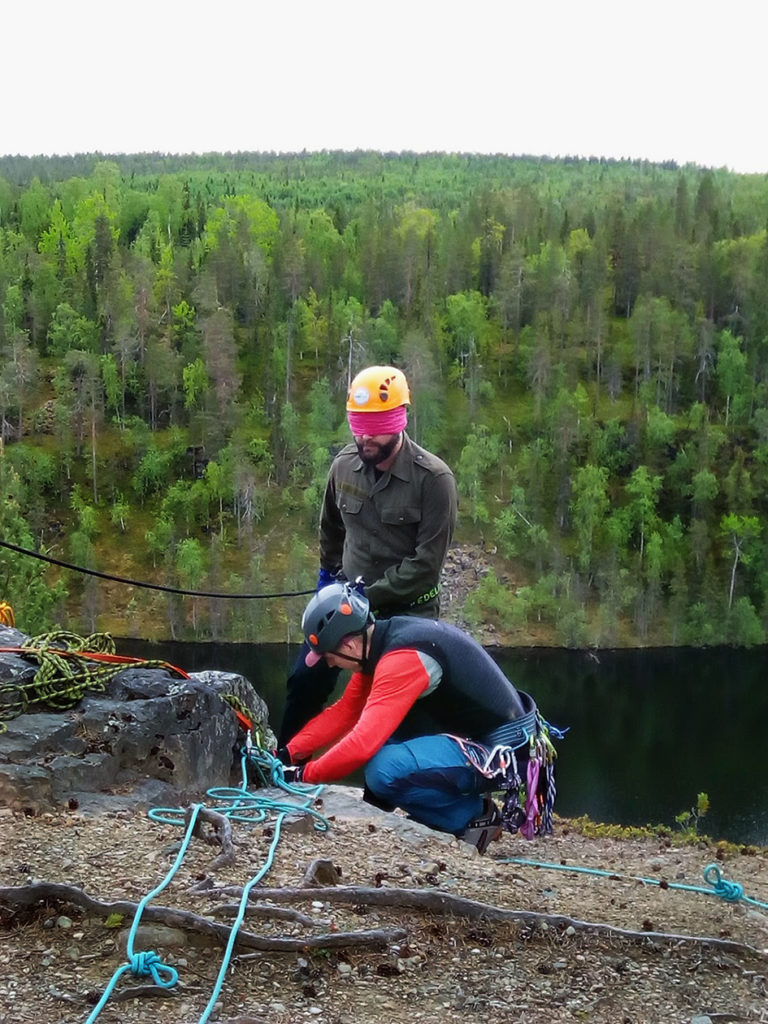 Jussi Tupasela varmistaa vielä Petri Markkasen kiinnitysköydet ennen laskua. Miehet ovat aivan jyrkänteen reunalla. Taustalla puiden peittämä vaara sekä sen edessä oleva vesistö. 