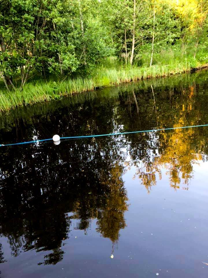 Tyyni lammen pinta jota halkoo kelluva kysy jossa on vaalea pallo merkkinä köyden päässä. Lammen takana kasvaa metsää joka heijastuu lampeen. 